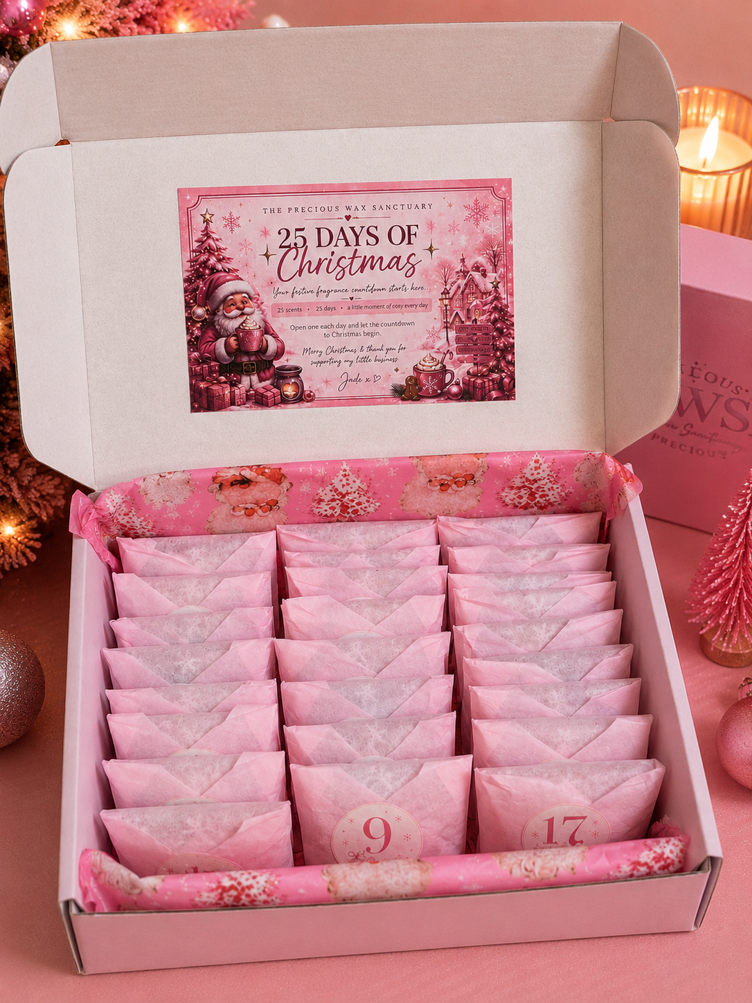 An open pink wax melt christmas advent calendar gift box containing 25 individually wrapped advent wax melts arranged in rows, with a festive Christmas-themed insert card inside the lid. The box is set on a pink surface with warm holiday lights and ornaments in the background.