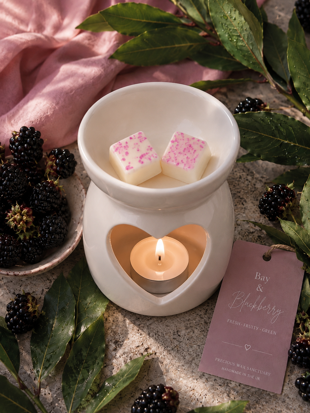A white ceramic wax warmer with a lit tea light candle inside, holding two small pink-sprinkled wax cubes. The scene is styled with blackberries, green leaves, a pink fabric cloth, and a mauve gift tag reading 'Buy & Blueberry'.