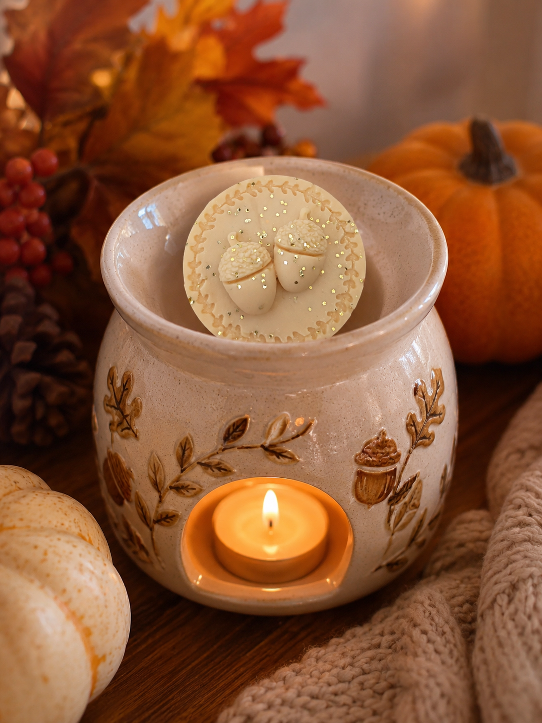 A ceramic wax warmer with a lit tea light candle inside and a round wax melt resting on the rim. The warmer is decorated with brown autumn leaf and acorn motifs.