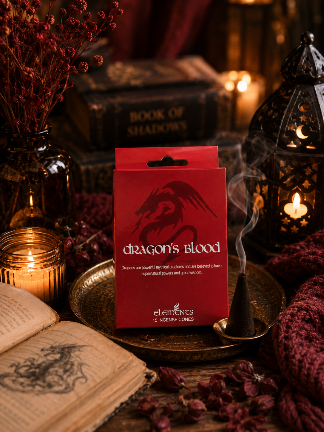 A red box of Elemences Dragon's Blood incense stands on a brass tray in a moody, mystical setup with burning candles, a lantern, dried flowers, and an open book.