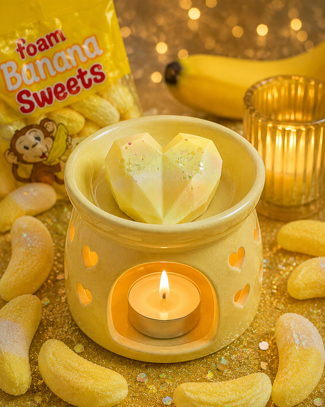 A yellow wax warmer with a lit tealight candle and a faceted heart-shaped wax melt on top, styled with banana-shaped candies, a yellow bag of foam banana sweets, and a glass candle holder on a glittery gold background.