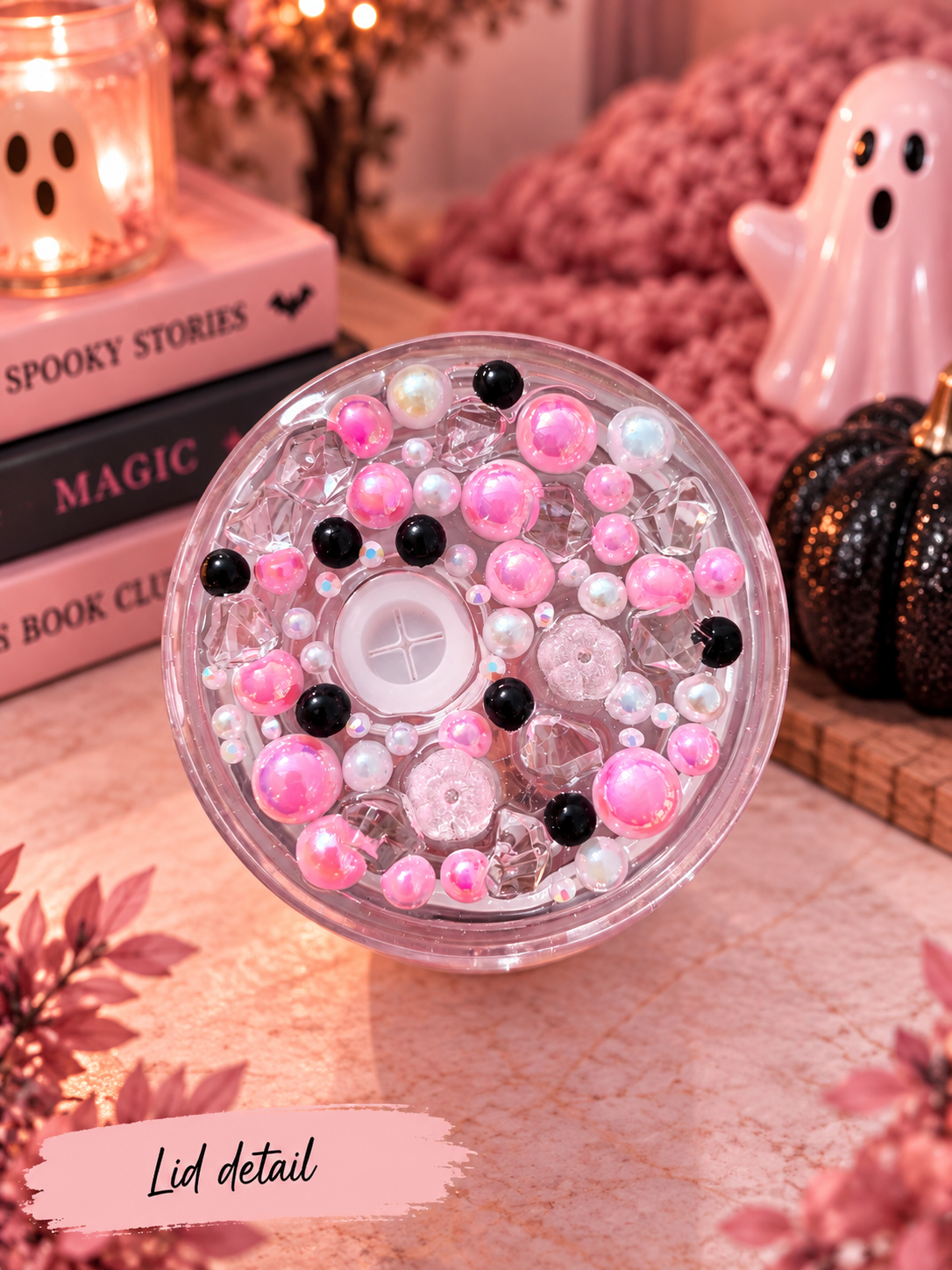 A round clear plastic container filled with pink, black, and iridescent beads, shown with a close-up label reading 'Lid detail' in the foreground. The scene is styled with a candle, books, a ghost figurine, and a small pumpkin in a warm, cozy setting.