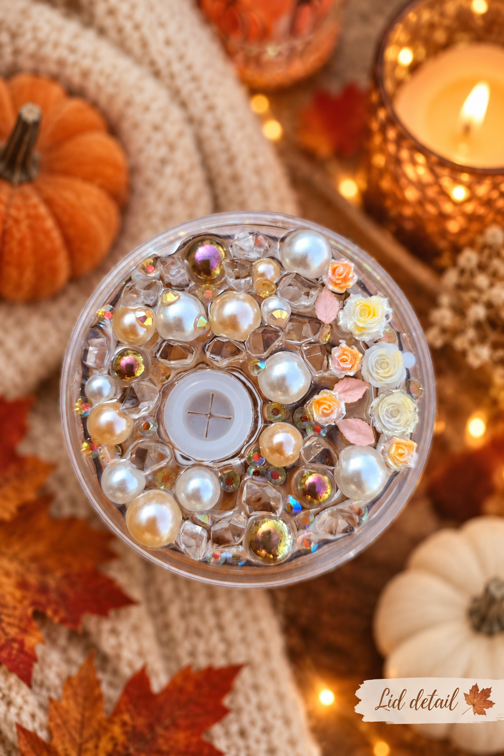 A round clear tumbler lid with decorative craft embellishments, including faux pearls, rhinestones, small rose-shaped beads, and iridescent beads, styled in an autumn setting with pumpkins, leaves, and a candle.