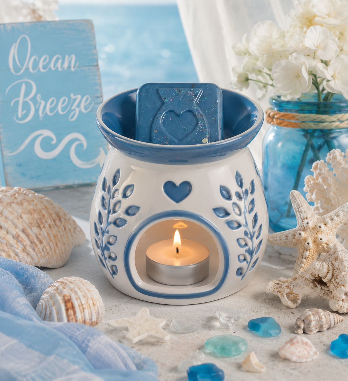 A ceramic wax warmer with blue leaf patterns and a wax melt inside, styled with seashells, a blue sign reading 'Ocean Breeze,' and a blue glass jar with white flowers on a beach-themed surface.