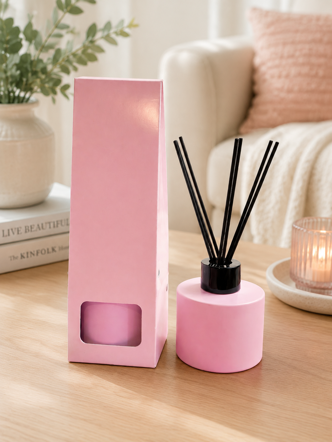 Pink reed diffuser set on a wooden table, featuring a cylindrical pink bottle with black reeds and a tall pink rectangular box with a window, styled in a cozy living room setting.