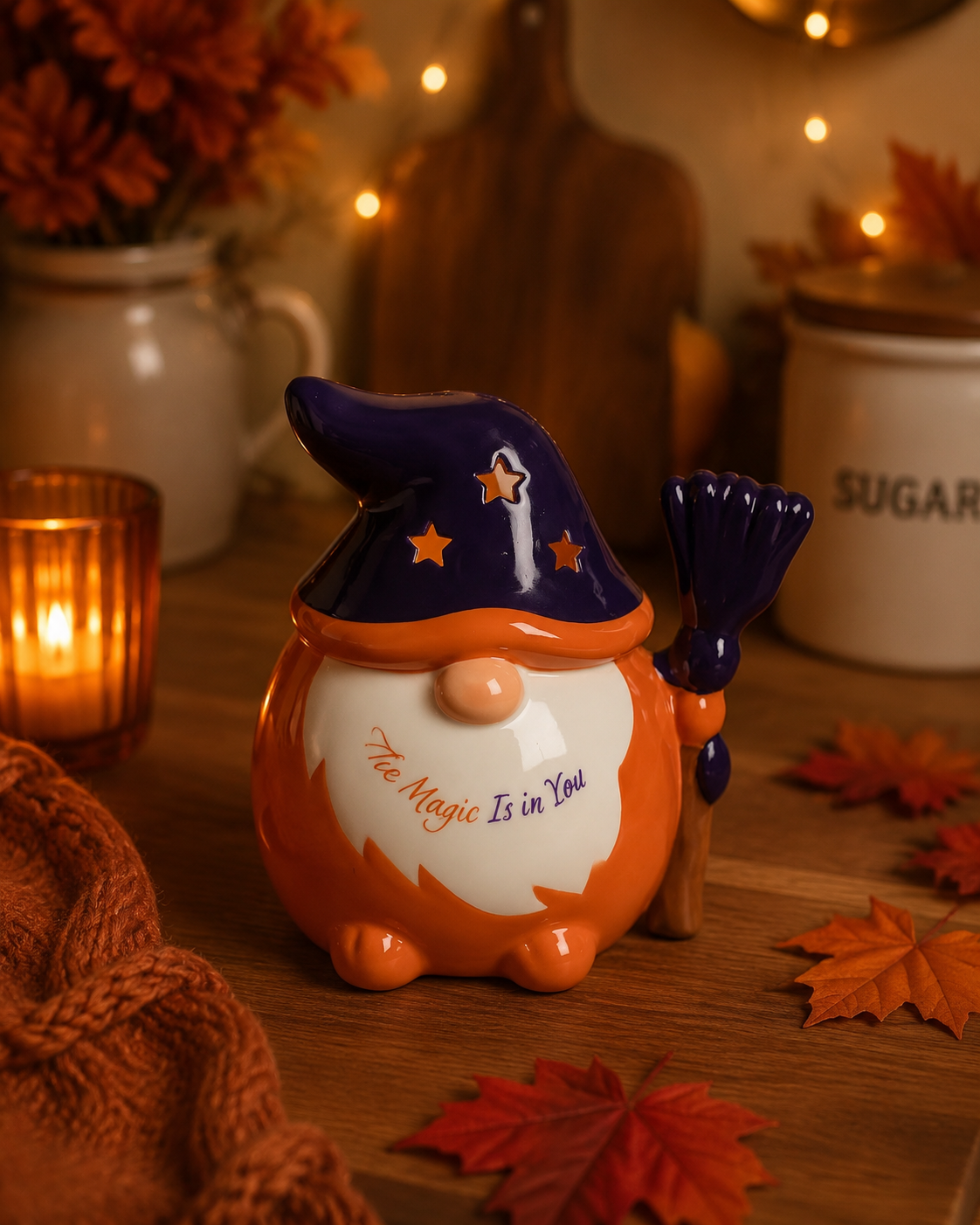 A ceramic witch gnome cookie jar with an orange body, dark blue hat, and a broom, displayed on a wooden table with autumn leaves, a candle, and warm background lights.