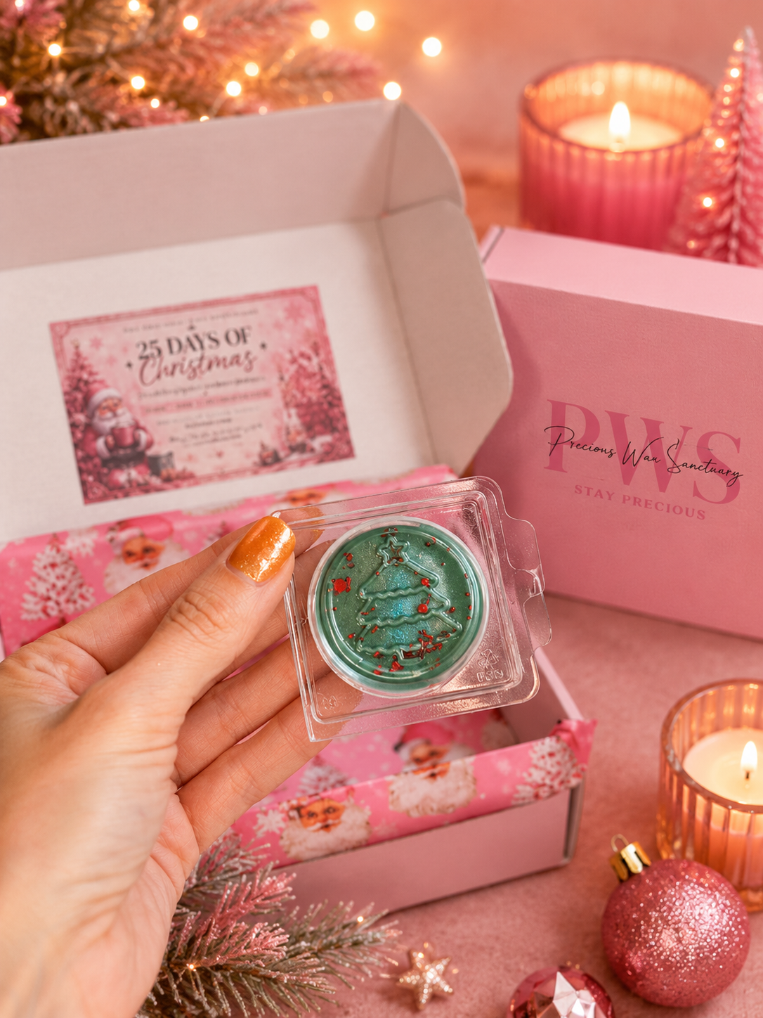 A hand with orange nail polish holds a small wax melt containing with glitter, set in front of an open pink holiday-themed box. The scene includes a pink branded box, candles, ornaments, and festive lights.