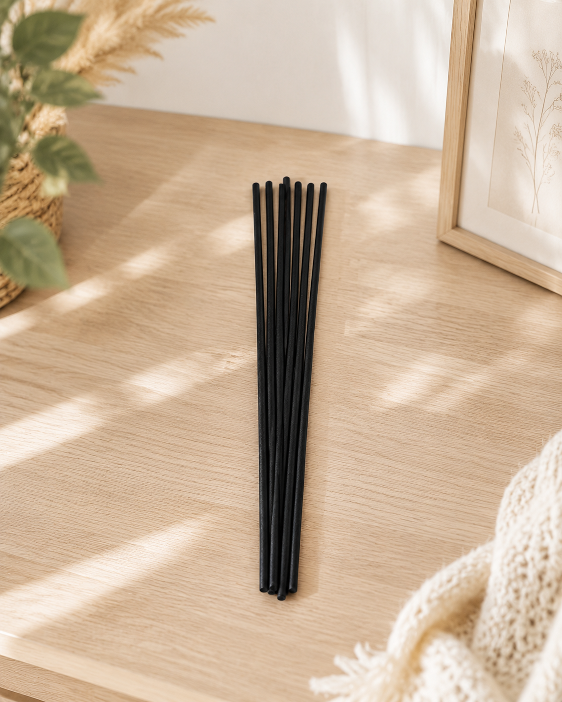 A set of 8 black reed diffuser sticks arranged in a vertical bundle on a light wood tabletop, styled with a potted plant, a framed print, and a cream knit blanket in soft natural light.
