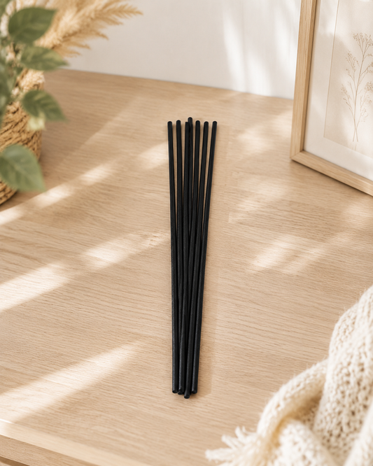 A set of 8 black reed diffuser sticks arranged in a vertical bundle on a light wood tabletop, styled with a potted plant, a framed print, and a cream knit blanket in soft natural light.