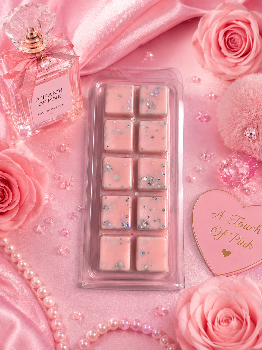 Pink wax melts in a clear plastic tray, styled on pink satin with pink roses, pearls, a perfume bottle labeled 'A Touch of Pink,' and a heart-shaped card reading 'A Touch of Pink.'.