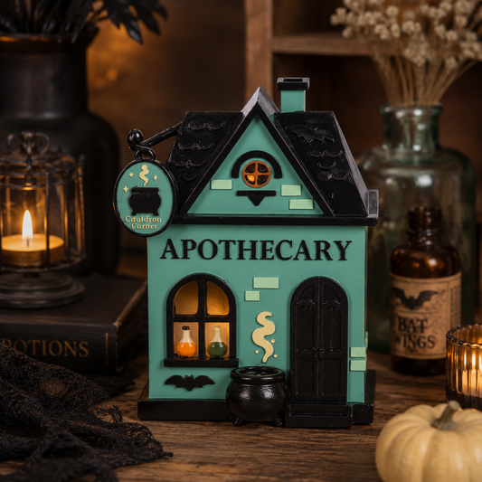 A decorative miniature house-shaped apothecary shop ornament with a teal and black design, surrounded by Halloween-themed props including a candle, potion bottles, black lace, and a small pumpkin on a wooden surface.
