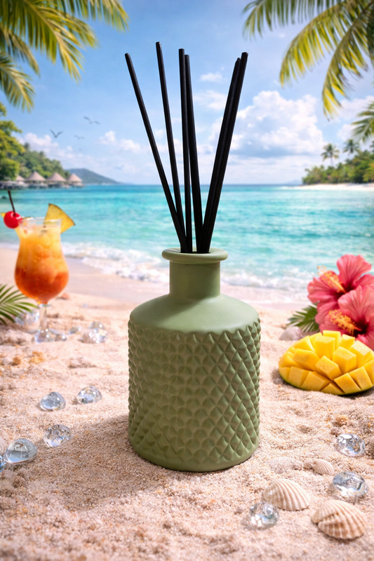 Green textured diffuser bottle with black sticks on a beach setting with a cocktail and flowers.