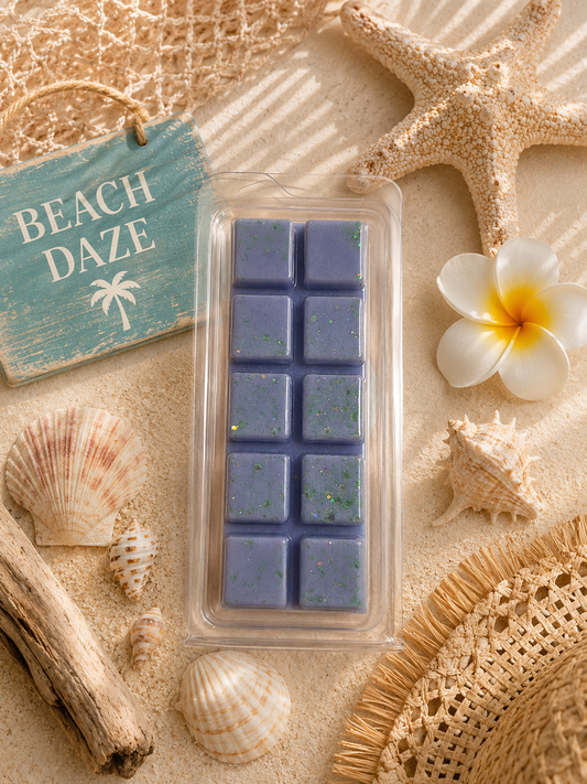 A rectangular wax melt in a clear clamshell package, placed on sand with beach-themed props including a starfish, seashells, driftwood, a woven hat, a plumeria flower, and a turquoise sign reading “BEACH DAZE.”.