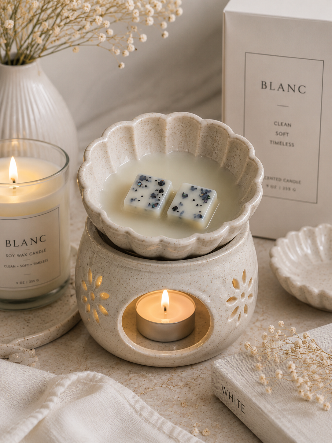 A ceramic wax warmer set with a scalloped bowl holding two wax melts, a lit tea light candle inside, and a matching candle jar and box labeled BLANC on a soft neutral surface with dried flowers and a small dish.