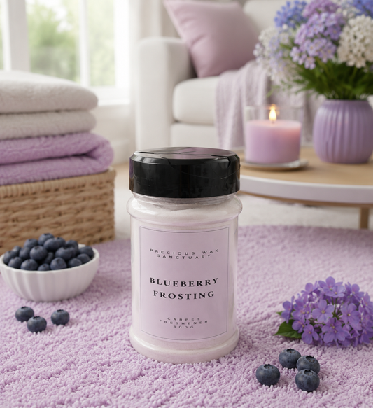 A jar of Blueberry Frosting carpet freshener sits on a lavender blanket in a cozy, softly lit room. The jar has a black lid and a pale pink label.