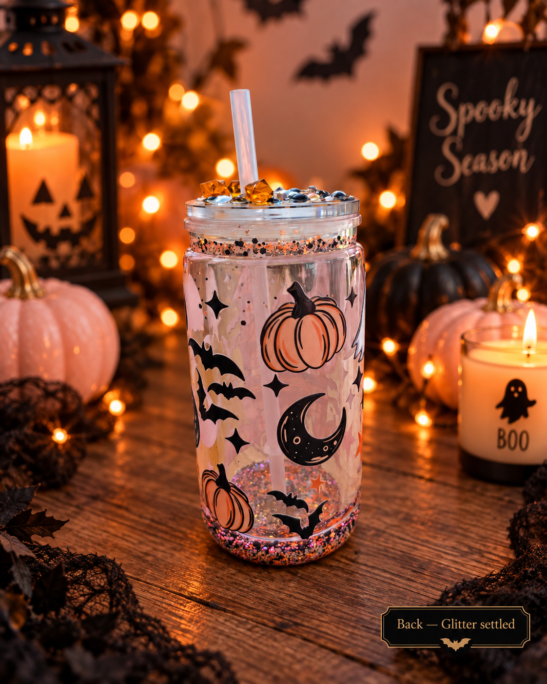 A clear glitter tumbler with a straw, decorated with pumpkins, bats, and a crescent moon, set on a wooden table surrounded by Halloween decorations including candles, pumpkins, and a sign that reads 'Spooky Season'.