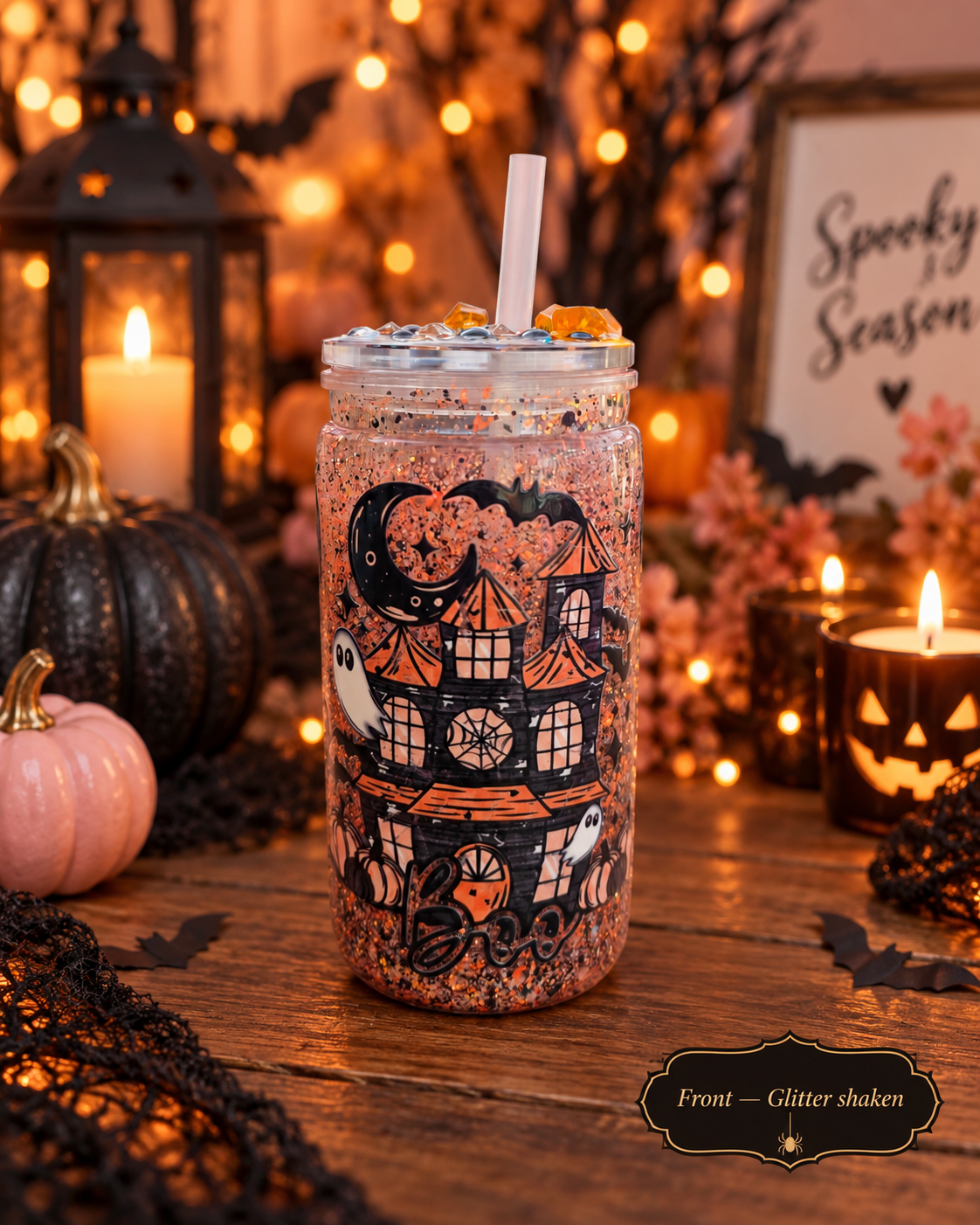 A glitter-shaken Halloween-themed mason jar tumbler with a clear lid and straw, decorated with a haunted house, ghosts, and bats. The tumbler is styled on a wooden table with warm string lights, candles, a small pink pumpkin, and black lace in a cozy autumn setting.