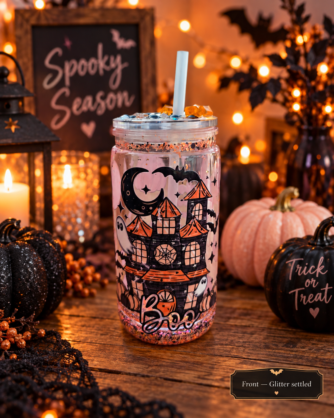 A clear glitter tumbler with a Halloween haunted-house design and a white straw, set on a rustic wooden table surrounded by fall decorations including black pumpkins, a candle, and a sign that reads 'Spooky Season'.