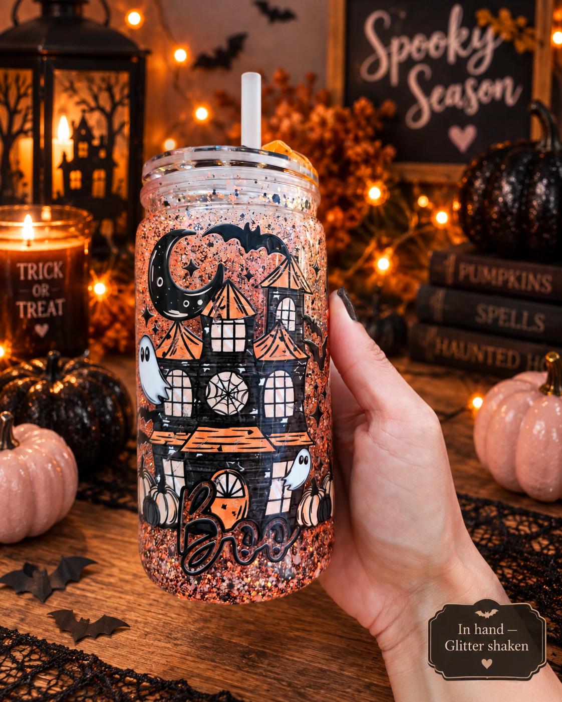 A hand holds a glittery mason jar tumbler with a white straw, decorated with a spooky haunted house, ghosts, bats, and the word 'Boo' in black glitter. The background is a cozy, warmly lit Halloween-themed setting with candles, pumpkins, and a lantern.