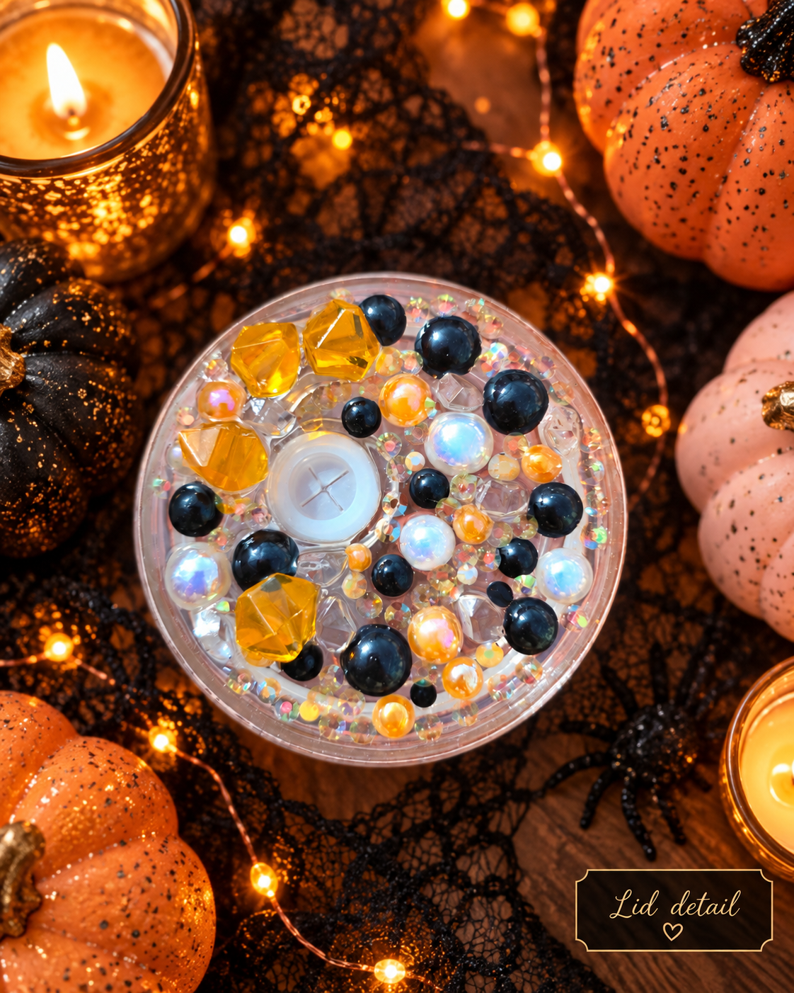 A round clear candle tin filled with decorative craft beads, rhinestones, and a white wax center, photographed from above on a dark Halloween-themed surface with black lace, string lights, pumpkins, and candles. A small label in the corner reads 'Lid detail'.