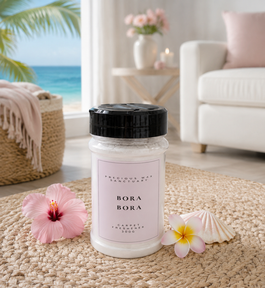 A jar of Bora Bora carpet freshener by Precious Bay Sanctuary sits on a woven jute rug in a beautifully styled beach-themed living room. The jar has a black lid and a pale pink label.
