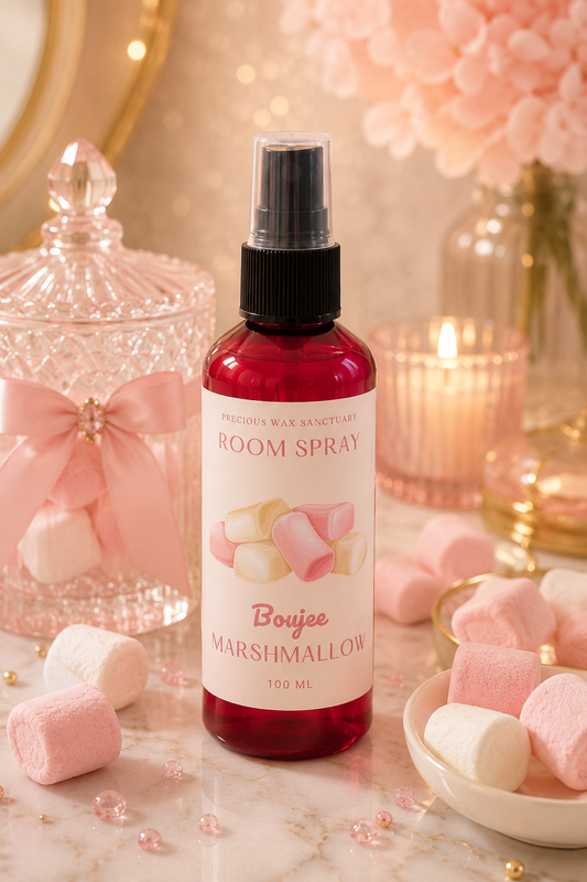 A bottle of room spray from Pretty Wax Sanctuary, scented with Boujee Marshmallow, displayed on a marble surface surrounded by pink marshmallows, a glass jar, and a lit candle.