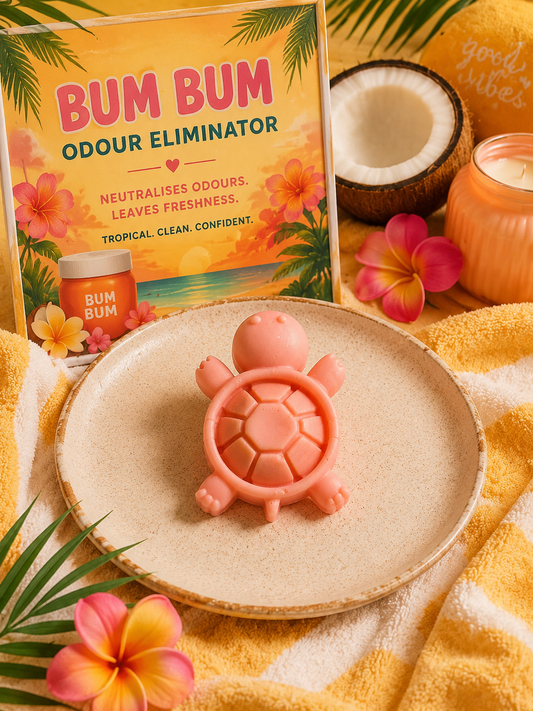 A pink turtle-shaped odour eliminator wax melt sits on a round beige plate, styled with a tropical beach theme including a 'Bum Bum' product box, a half coconut, pink flowers, a candle, and a yellow towel.