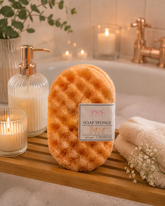 A PWS Bath & Body soap sponge in a waffle texture, displayed on a wooden bath caddy in a warm, relaxing bathroom setting with a towel, candles, and a glass bottle.