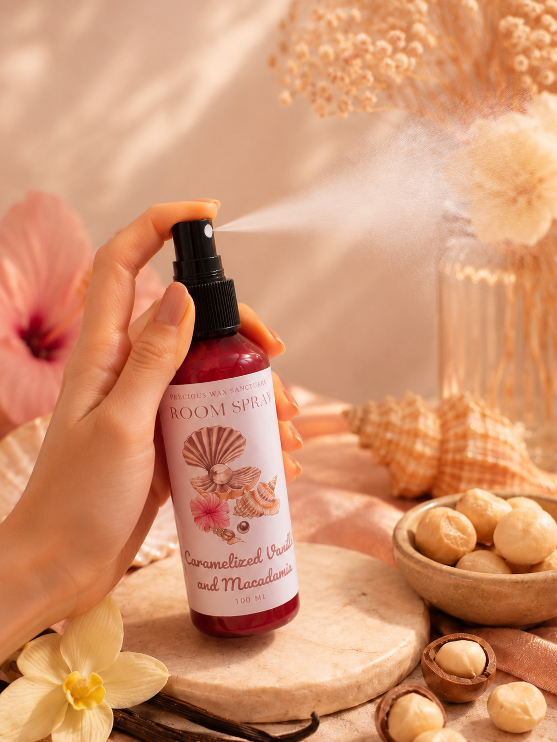 A hand holds a room spray bottle with a black spray nozzle, releasing a fine mist. The bottle has a white label with seashell illustrations and reads 'ROOM SPRAY' and 'Caramelized Vanilla and Macadamia'.