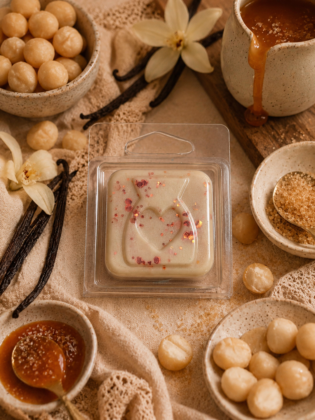Heart-shaped wax melt surrounded by vanilla beans, macadamia nuts, and a small pot of caramel on a textured surface.