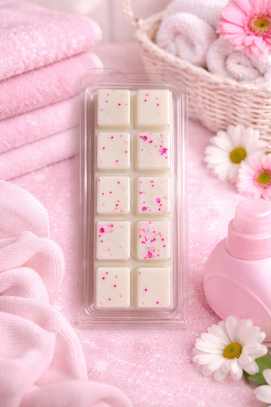 Pack of white wax melts with pink specks in a clear plastic case on a pink surface with flowers and towels.