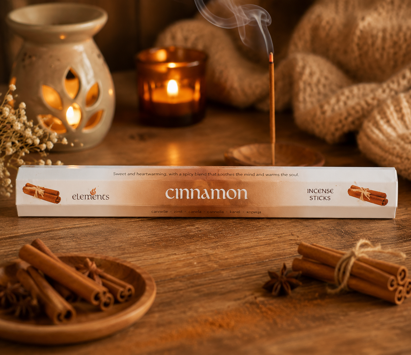 A box of Elements Cinnamon Incense Sticks lies on a rustic wooden table, surrounded by cinnamon sticks, star anise, a lit candle, and a ceramic diffuser in the background.