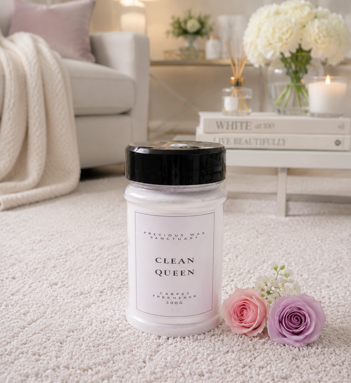 A jar of carpet freshener labeled 'CLEAN QUEEN' by Precious Wax Sanctuary sits on a white carpet in a softly lit living room. The jar has a white body and a black lid, with pink and lavender roses placed beside it.