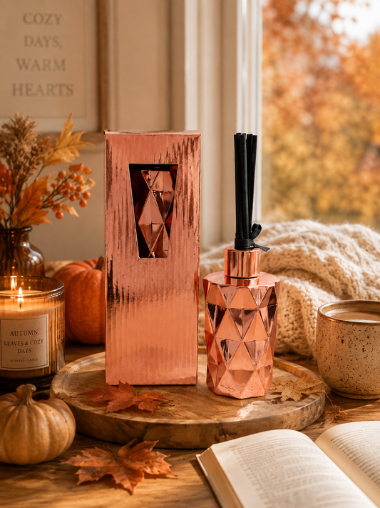 A faceted copper-toned reed diffuser bottle with a black reed stick stands beside its matching rectangular box on a wooden tray, styled in a cozy autumn scene with a candle, pumpkin, open book, mug, and window view.