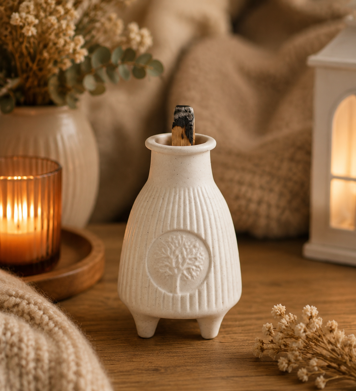 A small white ceramic incense holder with a ribbed texture and embossed tree motif, shown on a wooden surface with dried flowers, a lit candle, and a cozy knitted blanket in a warm indoor setting.