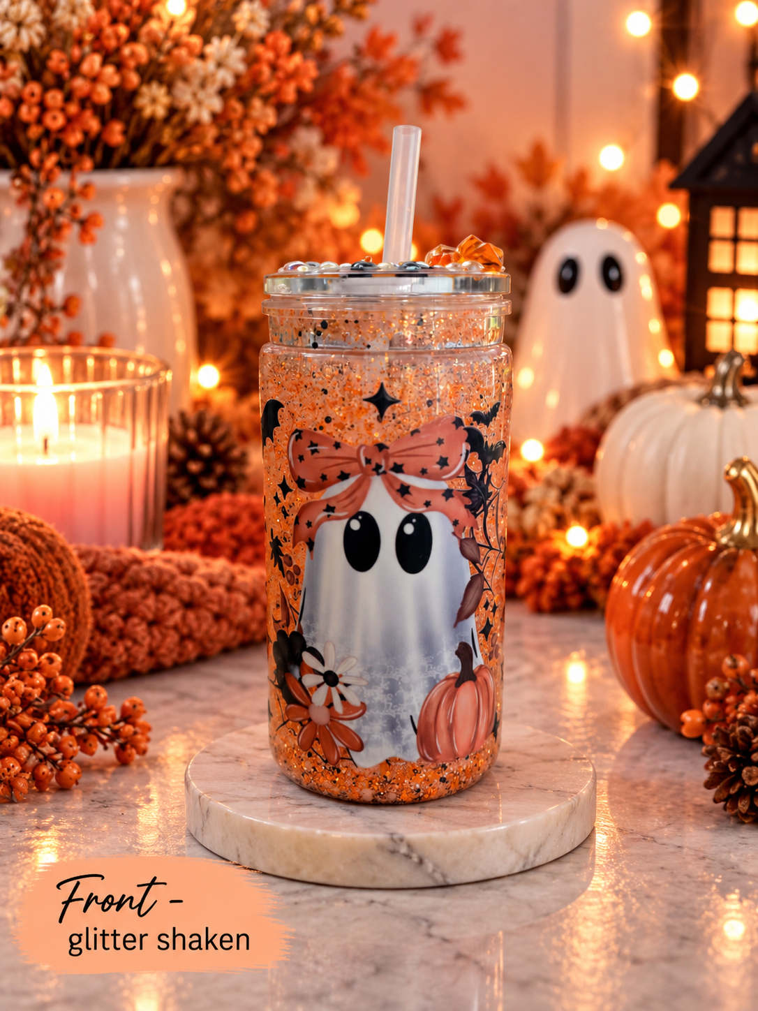 A clear tumbler with a lid and straw, filled with orange glitter and decorated with a ghost and pumpkin illustration, set on a marble coaster in an autumn-themed setting with pumpkins, candles, and string lights.