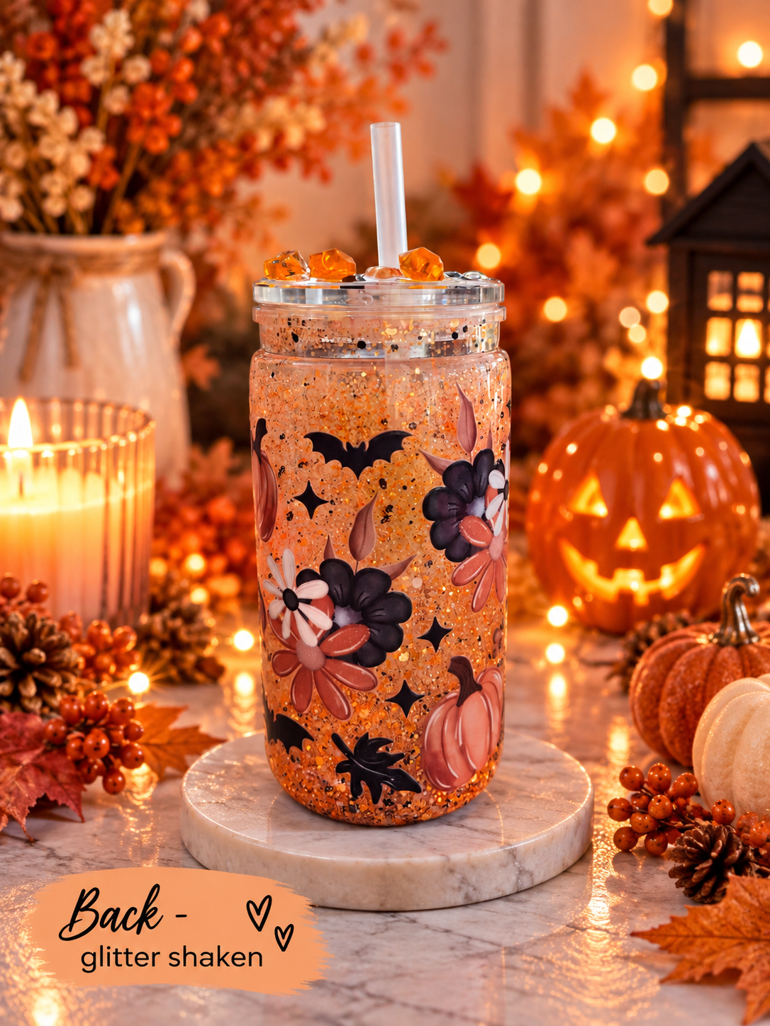 A glittery orange mason jar tumbler with a clear lid and straw, decorated with black bats, purple flowers, and pumpkins, set on a marble coaster in a warm autumn-themed setting with pumpkins, candles, and string lights.