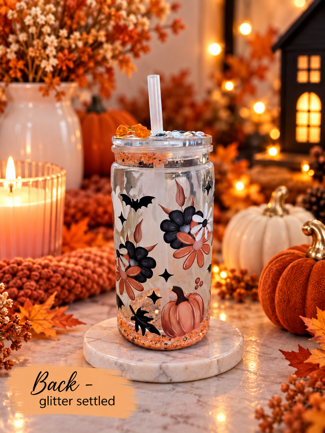 A clear glitter-settled mason jar tumbler with a clear lid and straw, decorated with black bats, orange pumpkins, and floral motifs, displayed on a marble coaster in a warm autumn-themed setting.
