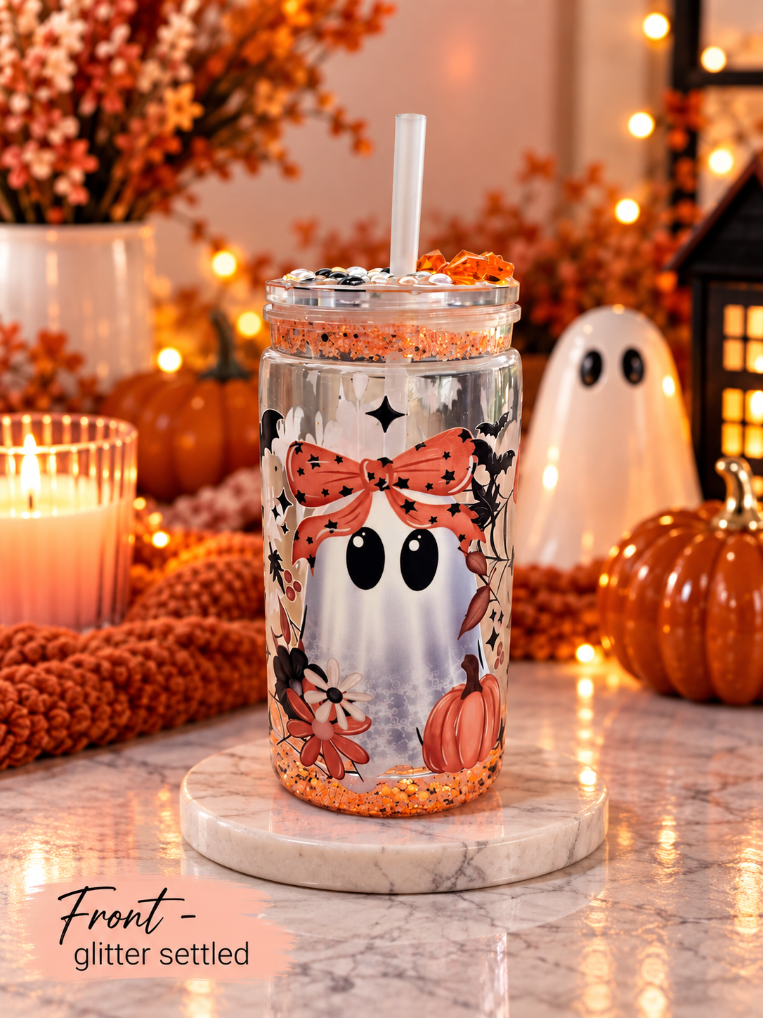 A clear tumbler with a Halloween-themed glitter settle design, featuring a white ghost face, orange bow, pumpkins, and bats, shown on a marble coaster with a straw in a cozy autumn setting.