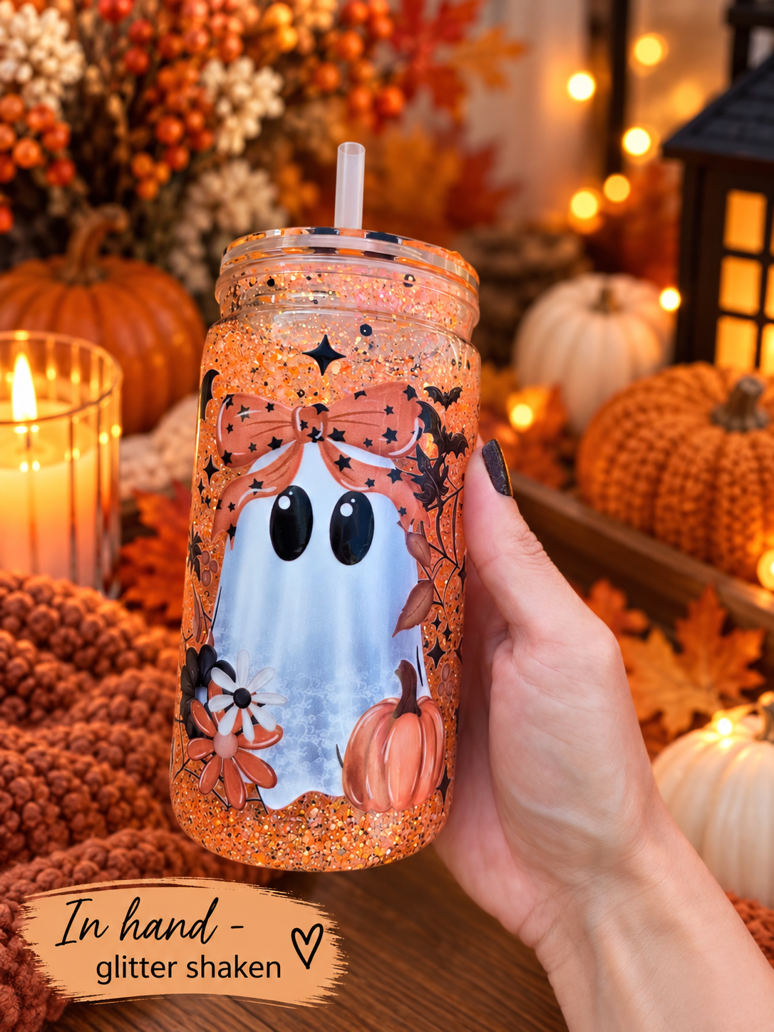 A hand holds a clear mason jar tumbler filled with orange glitter and decorated with a ghost, pumpkins, and autumn leaves. The background is a cozy, warm autumn scene with pumpkins, candles, and string lights.