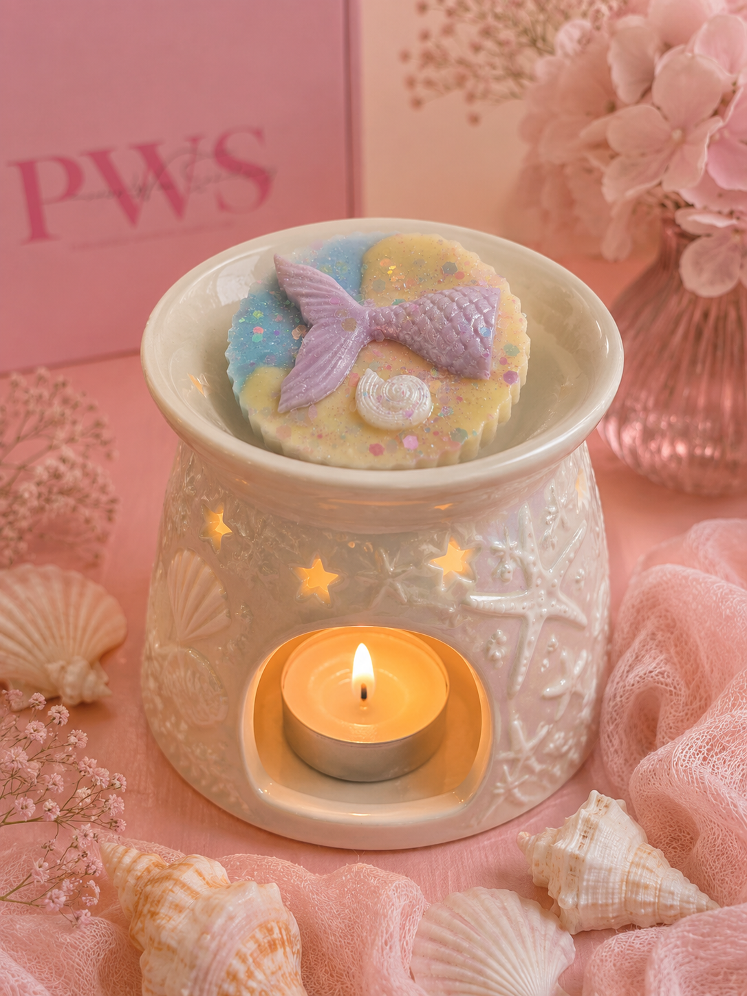 A ceramic wax warmer with embossed starfish and sea urchin details, lit with a small tealight candle inside. A round decorative soap or wax melt sits on top, featuring a lavender mermaid tail and pastel blue and yellow background with tiny pink and white accents.