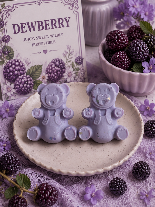 Two lavender-colored bear-shaped wax melts displayed on a speckled ceramic plate, with a 'DEWBERRY' card, fresh blackberries, and purple flowers in the background.