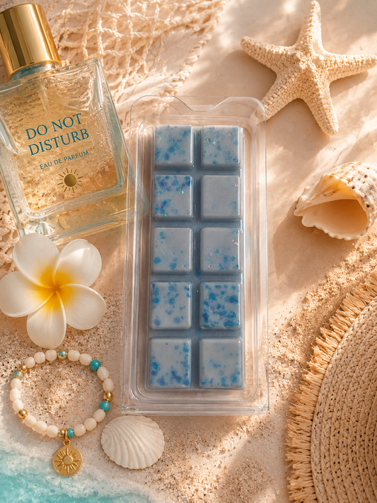 A packaged wax melt or scented melt bar set in a clear clamshell package, placed on a sandy beach-style surface with a starfish, seashell, bracelet, flower, and a perfume bottle nearby. The product itself is light blue with small darker blue speckles.