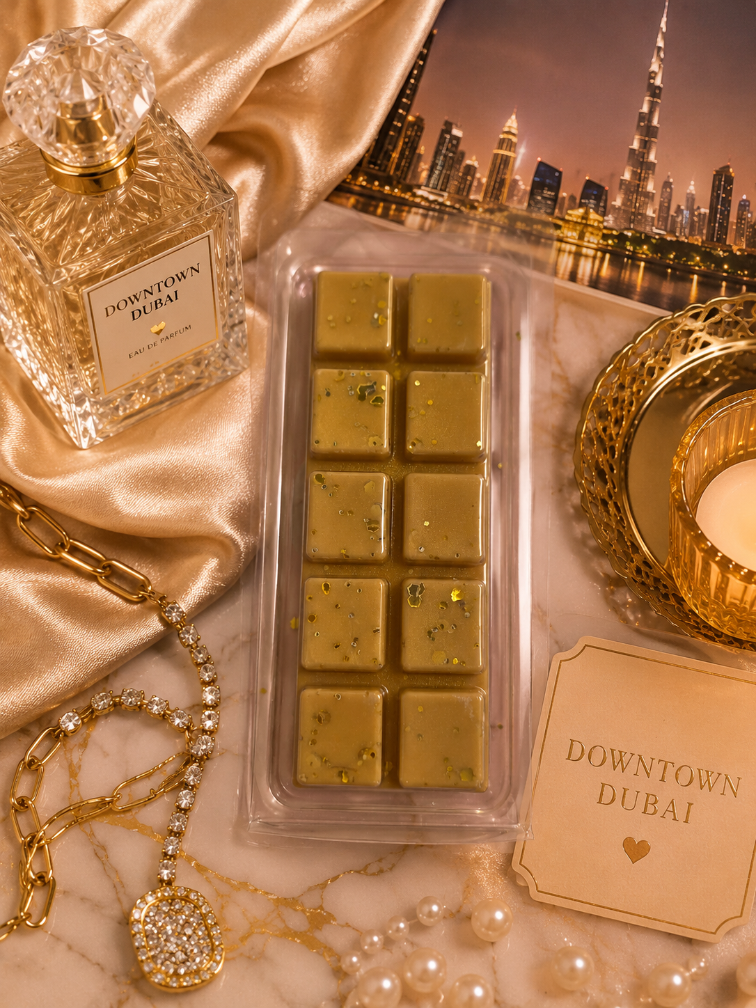 A clear plastic tray holding wax melts with tiny gold flecks, styled on a marble surface with champagne silk, a perfume bottle labeled Downtown Dubai, a gold candle holder, pearls, and a Downtown Dubai card.
