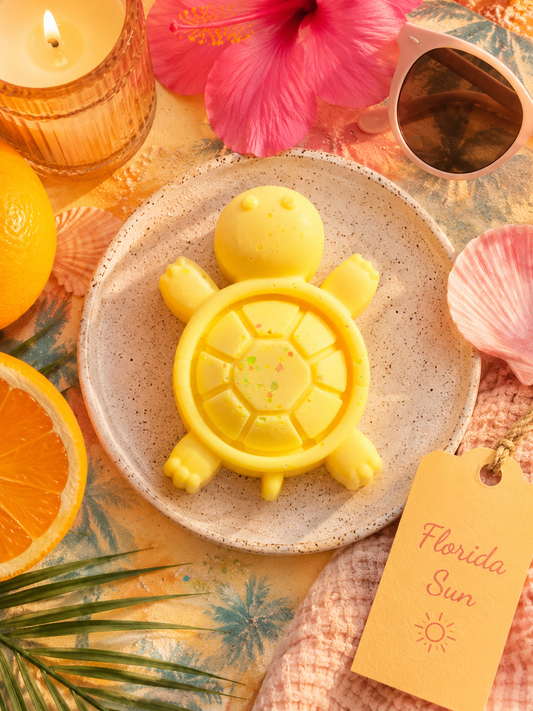 A yellow turtle-shaped wax melt sits on a speckled ceramic plate, surrounded by tropical props including a pink hibiscus flower, sunglasses, an orange slice, a candle, a pink seashell, and a yellow tag reading 'Florida Sun'.