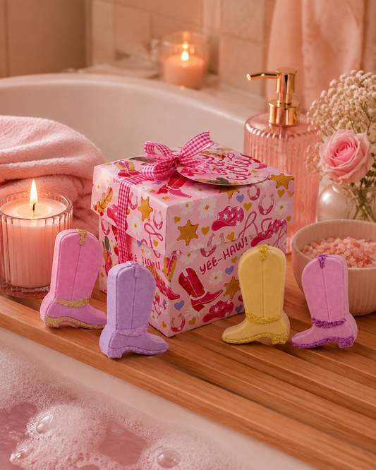 A pink and white cowboy boot themed gift box with a pink gingham bow sits on a wooden bath tray beside four boot-shaped bath bombs in pink, lavender, and yellow. The scene is styled in a warm, relaxing bathroom setting with a bathtub, lit candles, and pink towels.