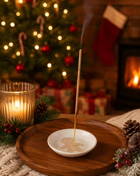 A single incense stick burns in a small white dish with a gingerbread man design, set on a round wooden tray. The scene is decorated for Christmas with a glowing candle, pinecones, evergreen branches, wrapped gifts, a Christmas tree with lights, and a fireplace in the background.
