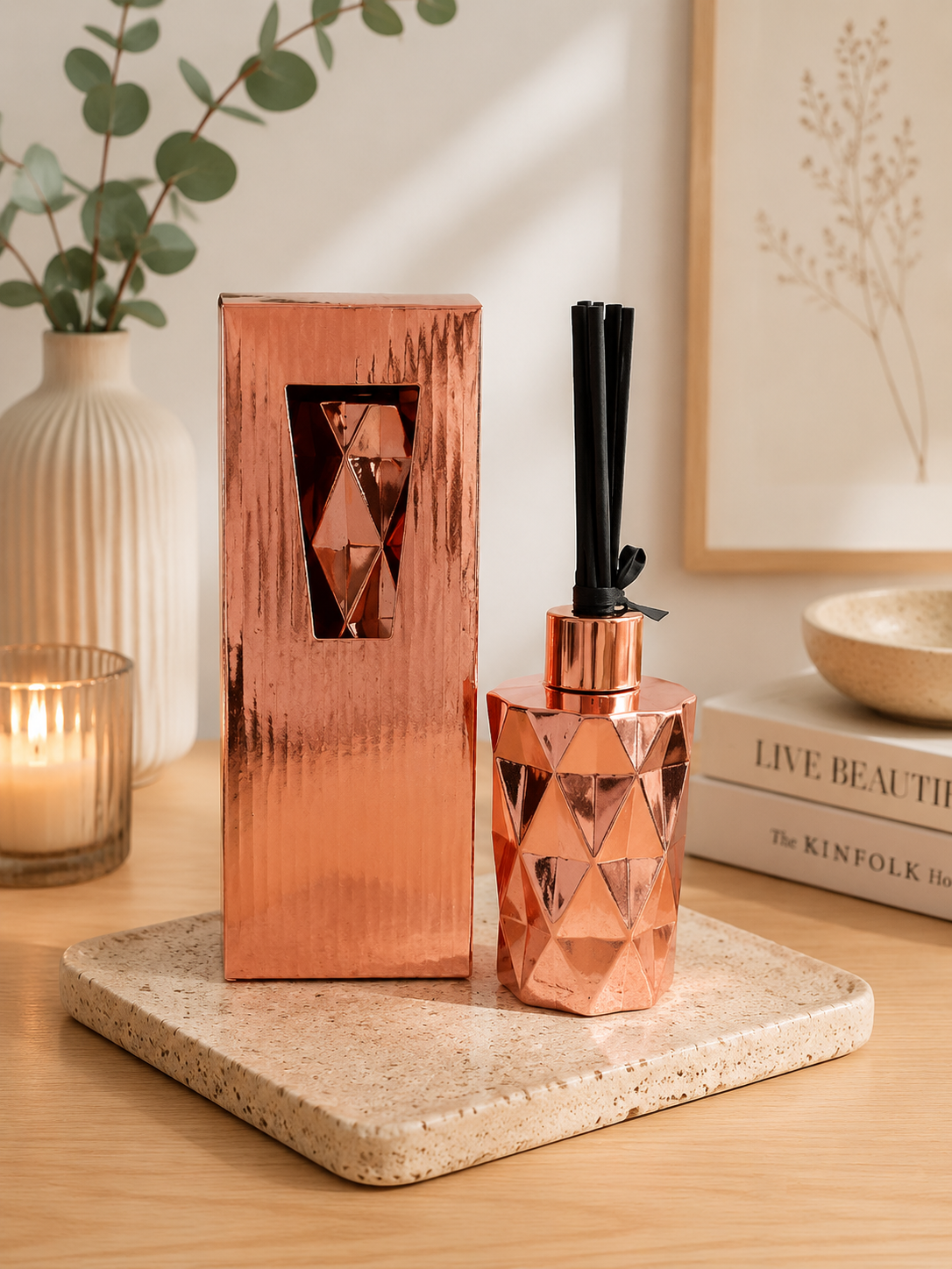 A faceted copper-toned reed diffuser bottle with black reeds stands beside its matching rectangular box on a stone tray, styled on a wooden table with a candle, vase, and framed art in the background.