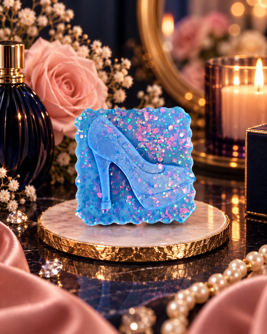 A square blue glitter wax melt shaped like a high-heeled shoe, displayed on a round marble coaster with pink roses, a dark perfume bottle, a lit candle, and pearls in the background.