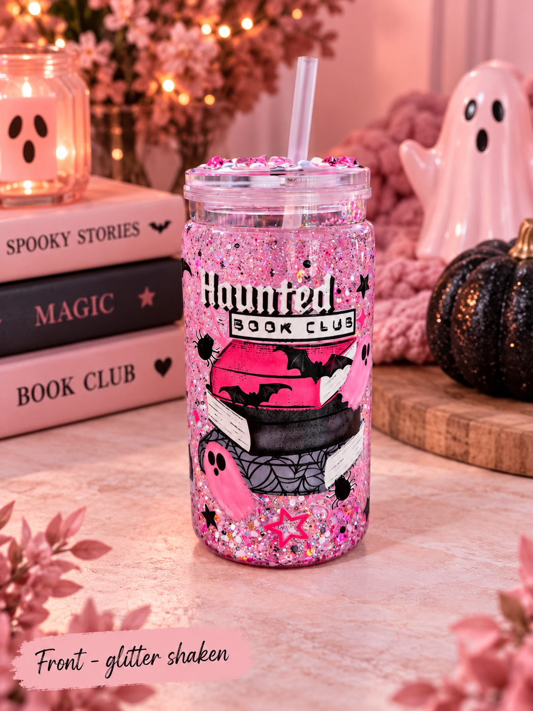 A pink glitter shaker tumbler with a straw, decorated with a 'Haunted Book Club' graphic, set on a tabletop with books, a ghost figurine, and a small pumpkin in a cozy, warm-toned setting.